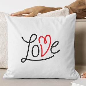 May include: White square pillow with the word "Love" in black script and a red heart-shaped "o". The pillow is on a light-colored surface.