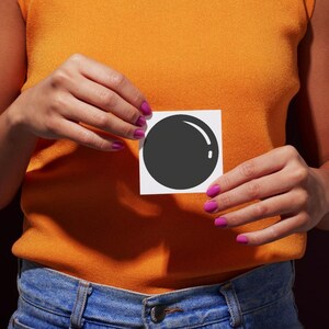 Könnte beinhalten: Eine Person hält ein weißes Quadrat mit einer schwarzen Kreisgrafik. Die Person trägt ein orangefarbenes Oberteil und blaue Jeans. Die Nägel sind leuchtend pink lackiert.