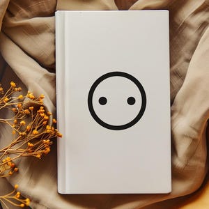 Puede incluir: Un libro blanco con un diseño circular negro con dos puntos negros. El libro está sobre una tela beige con flores amarillas secas en primer plano.