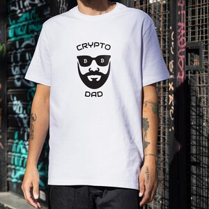 May include: White t-shirt with a black graphic of a bearded face wearing sunglasses, with the words "CRYPTO DAD" above and below the graphic. The shirt is short-sleeved and worn by a person.