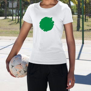May include: White crew neck t-shirt with a large green scribble design. The person is holding a soccer ball. The person is wearing black athletic pants. The background includes a chain-link fence and palm trees.