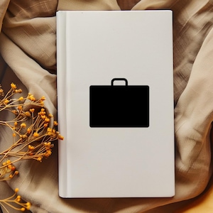 Op de afbeelding: Een wit boek met een zwart pictogram van een aktetas op de omslag. Het boek ligt op een beige stof, met gedroogde gele bloemen op de voorgrond. Het aktetas-pictogram is een eenvoudige, effen zwarte vorm.