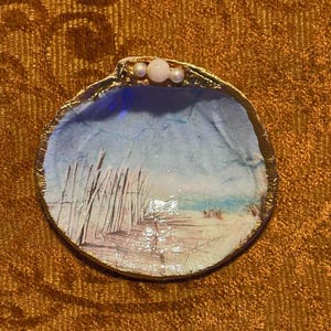 Puede incluir: Un cuenco decorativo de concha marina con una escena de playa pintada en su interior. El borde está bordeado en dorado y tres pequeñas perlas están adheridas a la parte superior. El interior presenta un cielo azul, arena y hierba alta.