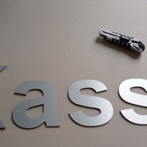 Puede incluir: Letras de metal plateado que deletrean la palabra "Kasse" sobre una superficie marr&oacute;n. Tambi&eacute;n se puede ver un cuchillo de uso general plateado en la imagen.