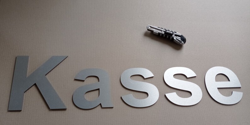 Puede incluir: Letras de metal plateado que deletrean la palabra "Kasse" sobre una superficie marr&oacute;n. Tambi&eacute;n se puede ver un cuchillo de uso general plateado en la imagen.
