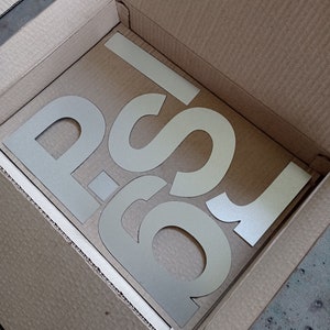 Puede incluir: Una caja de cart&oacute;n con las letras "P-S" recortadas en el cart&oacute;n. Las letras son de color beige claro.