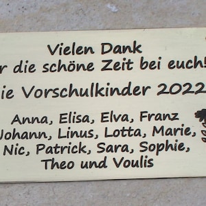May include: A gold-colored metal plaque with black text that reads "Vielen Dank f&uuml;r die sch&ouml;ne Zeit bei euch! Die Vorschulkinder 2022 Anna, Elisa, Elva, Franz Johann, Linus, Lotta, Marie, Nic, Patrick, Sara, Sophie, Theo und Voulis". The plaque has two small holes at the top and bottom.