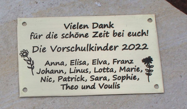 May include: A gold-colored metal plaque with black text that reads "Vielen Dank f&uuml;r die sch&ouml;ne Zeit bei euch! Die Vorschulkinder 2022 Anna, Elisa, Elva, Franz Johann, Linus, Lotta, Marie, Nic, Patrick, Sara, Sophie, Theo und Voulis". The plaque has two small holes at the top and bottom.