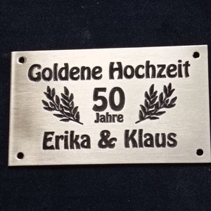May include: A silver metal plaque with black engraved text that reads "Goldene Hochzeit 50 Jahre Erika & Klaus".