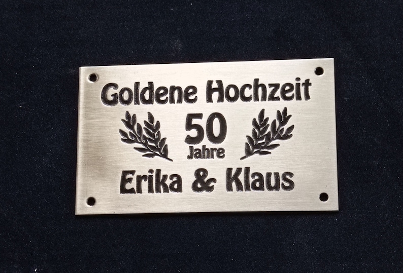May include: A silver metal plaque with black engraved text that reads "Goldene Hochzeit 50 Jahre Erika & Klaus".
