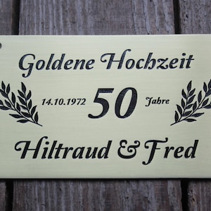 May include: A gold-colored metal plaque with black lettering. The plaque reads "Goldene Hochzeit" with the date "14.10.1972" and the number "50" followed by the word "Jahre". The plaque also reads "Hiltraud & Fred".
