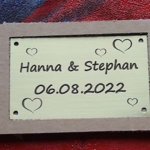 May include: A yellow metal plaque with the text "Hanna & Stephan" and the date "06.08.2022" engraved on it. The plaque is surrounded by a brown cardboard frame and has several small heart shapes engraved around the text.