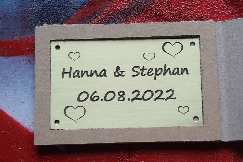 May include: A yellow metal plaque with the text "Hanna & Stephan" and the date "06.08.2022" engraved on it. The plaque is surrounded by a brown cardboard frame and has several small heart shapes engraved around the text.