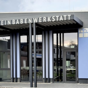 Puede incluir: Un edificio gris con un letrero que dice "Rheinbabenwerkstatt". El edificio tiene una fachada de vidrio con una pared azul en el lado derecho. Hay un logotipo en la pared azul que parece una cruz con un c&iacute;rculo alrededor.