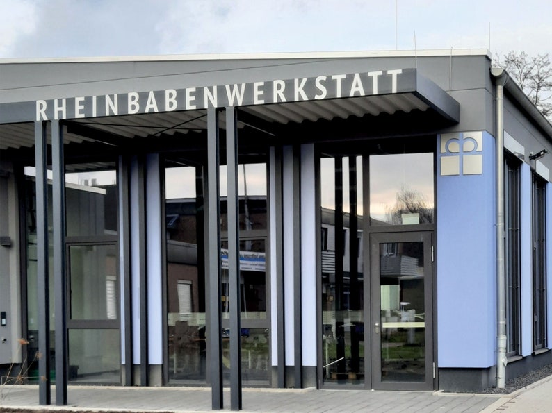 Puede incluir: Un edificio gris con un letrero que dice "Rheinbabenwerkstatt". El edificio tiene una fachada de vidrio con una pared azul en el lado derecho. Hay un logotipo en la pared azul que parece una cruz con un c&iacute;rculo alrededor.
