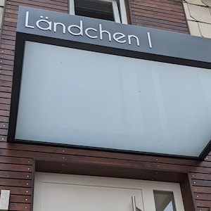 Puede incluir: Un toldo de vidrio con marco negro y el texto "L&auml;ndchen I" en letras blancas. El toldo est&aacute; montado sobre una puerta de madera con una puerta blanca.