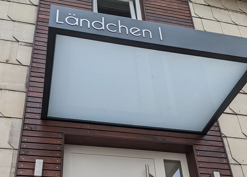 Puede incluir: Un toldo de vidrio con marco negro y el texto "L&auml;ndchen I" en letras blancas. El toldo est&aacute; montado sobre una puerta de madera con una puerta blanca.