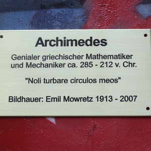 May include: A gold plaque with black text reads "Archimedes" and describes him as a "Genius Greek mathematician and mechanic ca. 285 - 212 v. Chr." The plaque also includes the quote "Noli turbare circulos meos" and the sculptor's name and dates "Bildhauer: Emil Mowretz 1913 - 2007".