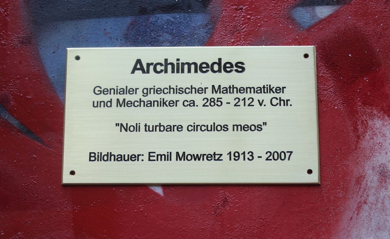 May include: A gold plaque with black text reads "Archimedes" and describes him as a "Genius Greek mathematician and mechanic ca. 285 - 212 v. Chr." The plaque also includes the quote "Noli turbare circulos meos" and the sculptor's name and dates "Bildhauer: Emil Mowretz 1913 - 2007".