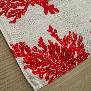 May include: A white fabric placemat with a red coral design. The placemat has a stitched edge.