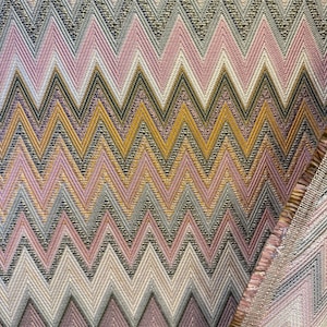 Puede incluir: Primer plano de una tela con un patrón de chevrón repetido. El diseño presenta una mezcla de colores, incluyendo rosa, dorado, gris y negro, creando una superficie texturizada y visualmente dinámica. Los bordes de la tela son visibles, mostrando la construcción del material.