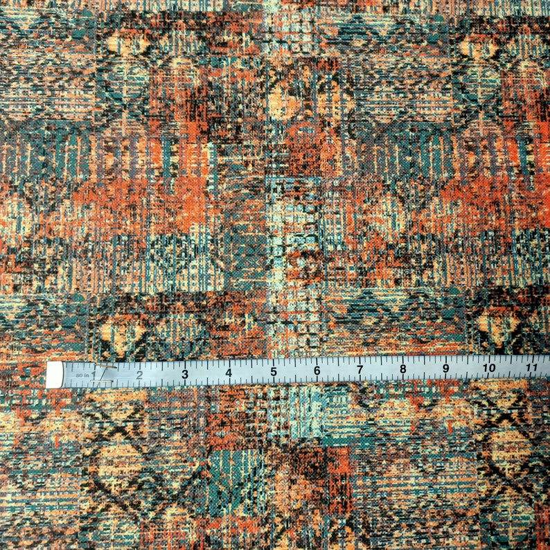 May include: A close-up of a fabric with a colorful, abstract pattern. The fabric is a mix of orange, blue, and brown colors, and has a textured, woven look. A ruler is placed on top of the fabric, showing the scale.