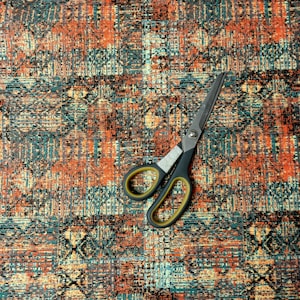 May include: A pair of silver scissors with black handles and yellow accents are lying on a multi-colored fabric with a geometric pattern.