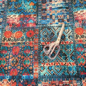 May include: A colorful fabric with a patchwork design featuring geometric patterns in shades of blue, red, orange, and white. A pair of silver scissors is placed on the fabric.