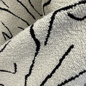 Könnte beinhalten: Nahaufnahme eines strukturierten, cremefarbenen Stoffes mit kräftigen schwarzen abstrakten Linien. Der Stoff scheint dick und weich zu sein, mit einer weichen, noppigen Textur. Das Design ist modern und grafisch, geeignet für die Inneneinrichtung.