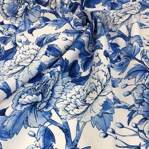 Op de afbeelding: Witte stof met een blauw bloemenpatroon. De bloemen zijn groot en hebben een aquareleffect. De stof is perfect voor knutselen, quilten of het maken van kleding.