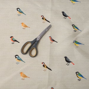 May include: A beige fabric with a repeating pattern of colorful birds. A pair of silver scissors is placed on the fabric.