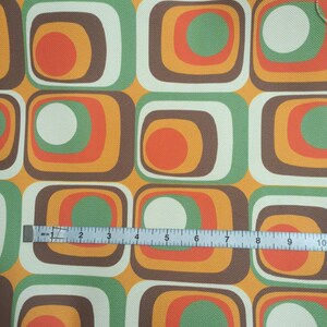 May include: A fabric with a repeating pattern of squares with concentric circles in shades of brown, orange, green, and white. A ruler is placed on the fabric to show the scale.