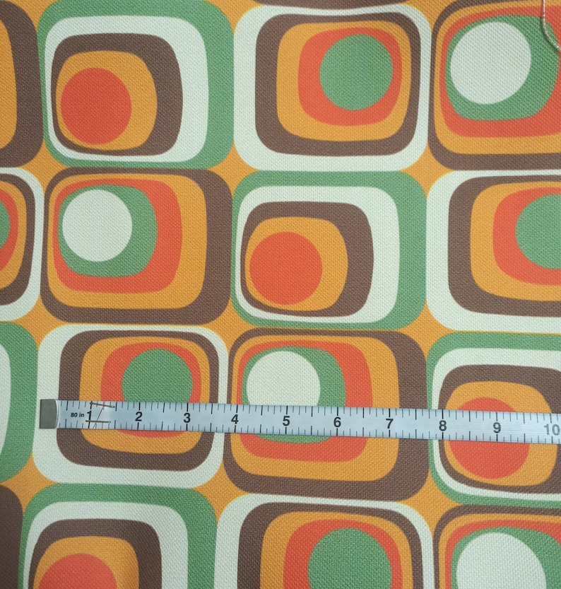 May include: A fabric with a repeating pattern of squares with concentric circles in shades of brown, orange, green, and white. A ruler is placed on the fabric to show the scale.