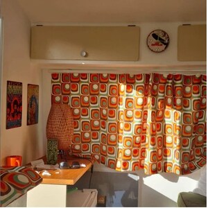 May include: A vintage camper interior with orange, brown, and white geometric patterned curtains. A wicker lamp sits on a wooden table with a mug and sunglasses.