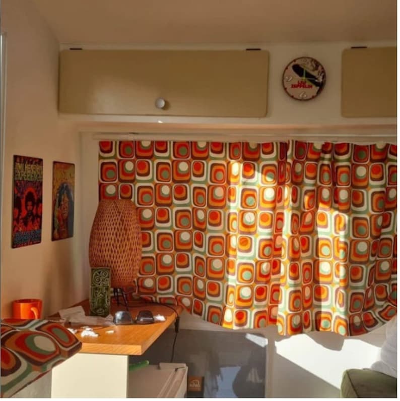 May include: A vintage camper interior with orange, brown, and white geometric patterned curtains. A wicker lamp sits on a wooden table with a mug and sunglasses.