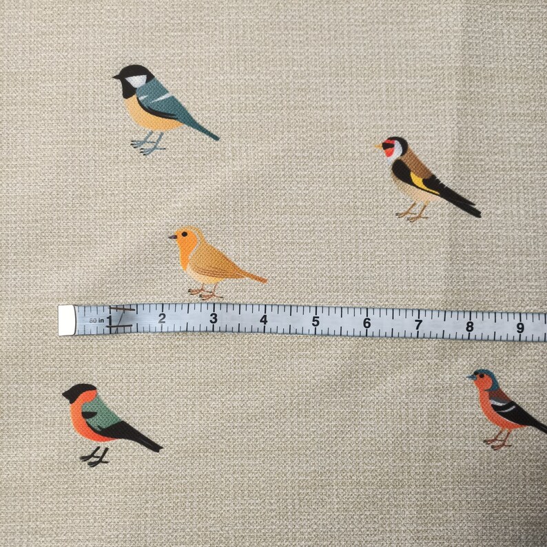 May include: A beige fabric with five embroidered birds. The birds are a blue and green bird, a yellow and red bird, an orange bird, a red and green bird, and a black and white bird. A ruler is placed below the birds.