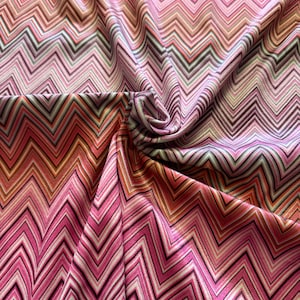 Könnte beinhalten: Nahaufnahme eines Stoffes mit einem lebendigen Zickzackmuster. Das Design zeigt ein sich wiederholendes Zickzack in Rosa-, Rot-, Orange- und Weißtönen. Der Stoff ist drapiert und zeigt die Textur und den Fluss des Materials. Ideal für Kleidung oder Heimdekoration.