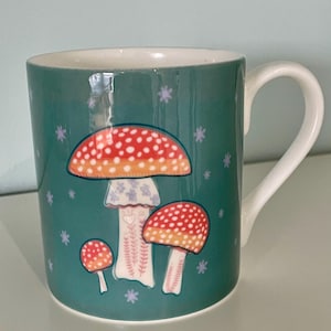 May include: A teal blue ceramic mug with a white handle features a whimsical design of three red and white mushrooms with orange accents on a teal blue background. The mushrooms have white dots and a white floral pattern.