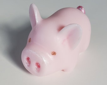 May include: A small, pink pig figurine with a rounded body, small ears, and a curled tail. The pig has a detailed snout with two nostrils and small, glittery accents on its face and tail. The figurine is set against a plain white background.