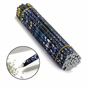 May include: A bundle of 50 black diamond painting pens with a blue and gold diamond pattern. The pens are wrapped together with a gold rubber band. A close-up of one pen shows a clear tip with a small diamond-shaped opening.