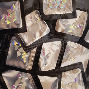 May include: Assortment of small clear rhinestone embellishments packaged in individual clear plastic bags with black borders. The bags are stacked on top of each other. The bags are labeled with the number "8".