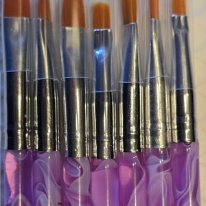 May include: A set of eight paint brushes with orange bristles and silver ferrules. The handles are purple with a white marbled design. The brushes are of various sizes and shapes, suitable for detailed painting work.