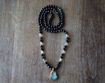 May include: A black beaded necklace with a large, iridescent blue gemstone pendant. The necklace is made with black and clear beads and has a gold accent on the pendant.
