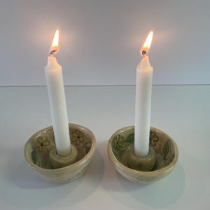 以下が含まれることがあります： 2つの白いテーパーキャンドルが、セラミック製のキャンドルホルダーに灯されています。キャンドルホルダーはベージュ色で、緑と黄色の花柄のデザインです。炎は明るい黄色とオレンジ色です。
