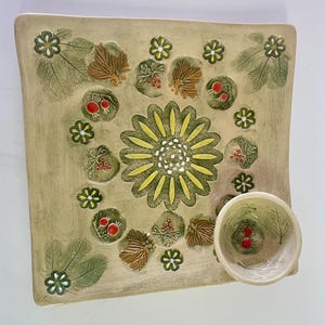 Peut inclure: Un plat de service carré en céramique avec un petit bol assorti. Le plat est beige avec un motif central de fleur jaune et verte, entouré de feuilles vertes, de feuilles brunes et de baies rouges. Le bol est sur le côté.