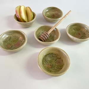 Könnte beinhalten: Satz von sechs kleinen, weißen Keramikschalen mit grünen und braunen Blumendesigns. Die Schalen sind in einem Kreis angeordnet, in der Mitte befindet sich ein Holzhonigrührer. Eine kleine Schale mit Apfelscheiben befindet sich im Hintergrund.