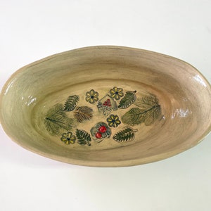 May include: A light beige oval ceramic serving dish with a hand-painted design of green leaves, yellow flowers, and red berries.