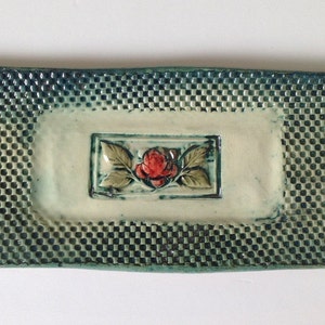 Puede incluir: Una bandeja rectangular de cerámica con un patrón de cuadros azules y verdes. La bandeja tiene un rectángulo blanco elevado en el centro con una rosa roja y hojas verdes.