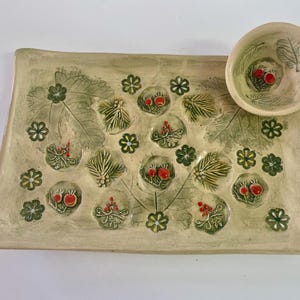 Könnte beinhalten: Ein rechteckiges Keramik-Serviertablett und eine kleine Schüssel. Das Set hat eine hellbeige Farbe mit grünen Blatt- und Beerenmotiven. Die Schüssel befindet sich in der oberen rechten Ecke des Tabletts. Das Tablett hat einen erhöhten Rand.