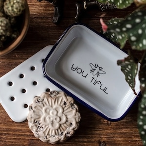 May include: White ceramic soap dish with a blue rim and the text "you tiful" with a bee graphic. The dish is sitting on a wooden surface with a white ceramic soap dish with holes and a white soap bar with a floral design.